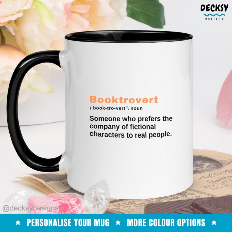 May include: White coffee mug with a black rim and handle. The mug has the text "Booktrovert 'book-tro-vert' noun Someone who prefers the company of fictional characters to real people." printed in orange and black.