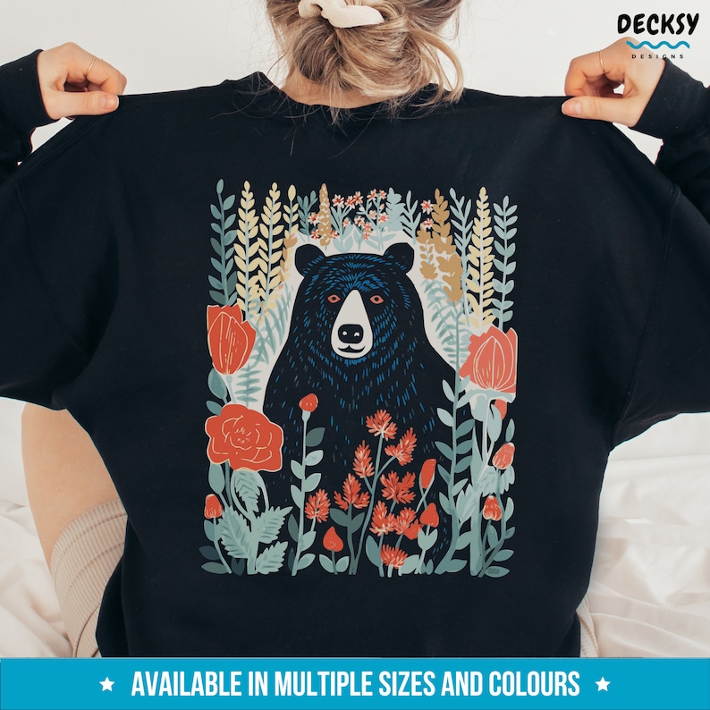 May include: Black sweatshirt with a graphic print of a black bear in a field of flowers. The flowers are red, orange, and yellow. The bear is surrounded by green leaves and stems.