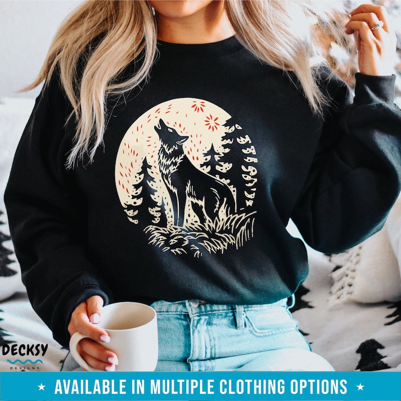 May include: Black sweatshirt with a graphic print of a wolf howling at the moon in a forest setting. The wolf is silhouetted against a yellow moon with red stars. The text "AVAILABLE IN MULTIPLE CLOTHING OPTIONS" is at the bottom of the image.