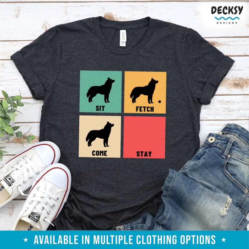 Op de afbeelding: Een donkergrijs T-shirt met een retro-ontwerp met een silhouet van een husky-hond in vier verschillende poses. De poses zijn gelabeld met "SIT", "FETCH", "COME" en "STAY".