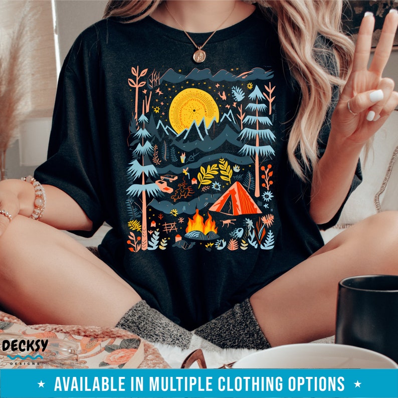 May include: A black t-shirt with a colorful illustration of a campsite scene. The design features a campfire, a tent, trees, and a full moon. The text "AVAILABLE IN MULTIPLE CLOTHING OPTIONS" is at the bottom of the image.