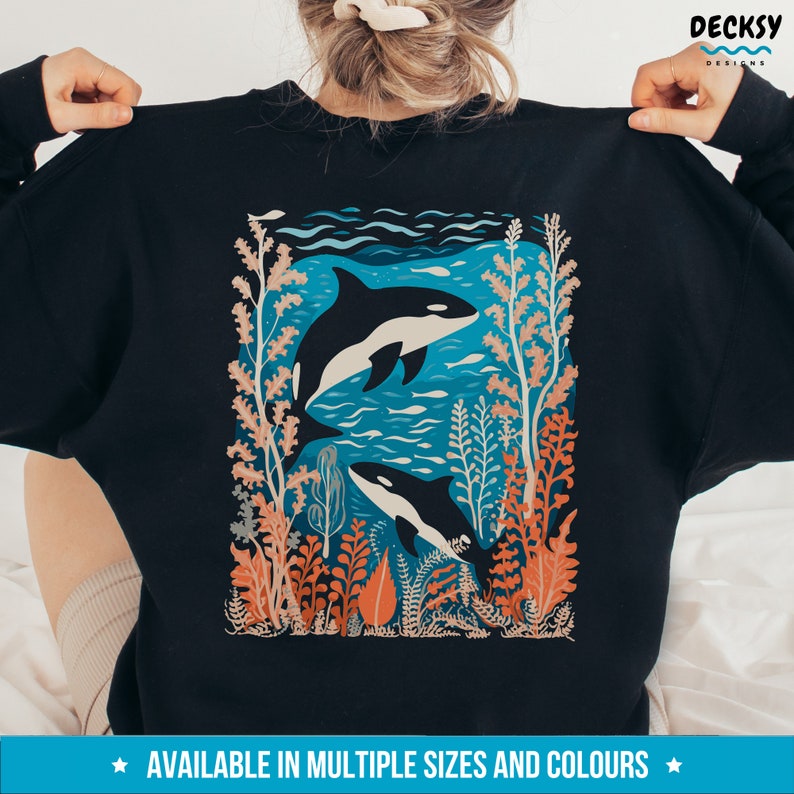 May include: Black sweatshirt with a graphic print of two orcas swimming in a blue ocean with coral and seaweed. The text "AVAILABLE IN MULTIPLE SIZES AND COLORS" is printed at the bottom of the image.