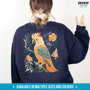 May include: A navy blue sweatshirt featuring a colourful illustration of a cockatoo bird in a floral design on the back. The design depicts a yellow and orange cockatoo with green and blue accents, surrounded by flowers and leaves.