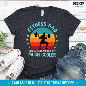 Puede incluir: Una camiseta gris oscuro con un gr&aacute;fico de estilo vintage que dice "Fitness Dad" con una silueta de una persona levantando pesas. El gr&aacute;fico tambi&eacute;n incluye una puesta de sol con un esquema de color rojo, naranja y amarillo. El texto debajo del gr&aacute;fico dice "Como un padre normal pero mucho m&aacute;s cool."