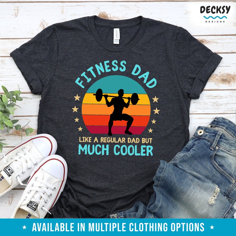 Puede incluir: Una camiseta gris oscuro con un gr&aacute;fico de estilo vintage que dice "Fitness Dad" con una silueta de una persona levantando pesas. El gr&aacute;fico tambi&eacute;n incluye una puesta de sol con un esquema de color rojo, naranja y amarillo. El texto debajo del gr&aacute;fico dice "Como un padre normal pero mucho m&aacute;s cool."