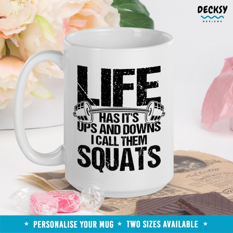May include: White ceramic mug with a black graphic that reads "Life has it's ups and downs I call them squats" with a barbell graphic above the text. The mug has a black handle and is sitting on a table with a brown surface.