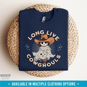 May include: Navy blue sweatshirt with a graphic featuring a cartoon ghost wearing a cowboy hat. The text reads "LONG LIVE COWGHOULS" in a vintage font. The design includes starburst and star accents. The bottom of the image states "AVAILABLE IN MULTIPLE CLOTHING OPTIONS".