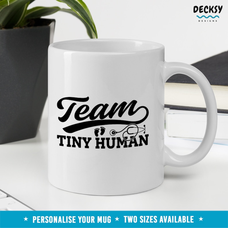 K&ouml;nnte beinhalten: Eine wei&szlig;e Keramiktasse mit einem schwarzen Design, auf dem "Team Tiny Human" mit einem Stethoskop und Fu&szlig;abdr&uuml;cken steht.