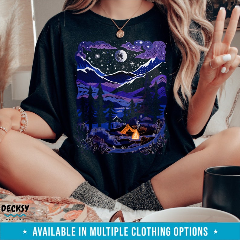 May include: A black t-shirt with a graphic of a purple mountain range with a full moon and stars. A campfire and a tent are in the foreground. The text "AVAILABLE IN MULTIPLE CLOTHING OPTIONS" is at the bottom of the image.