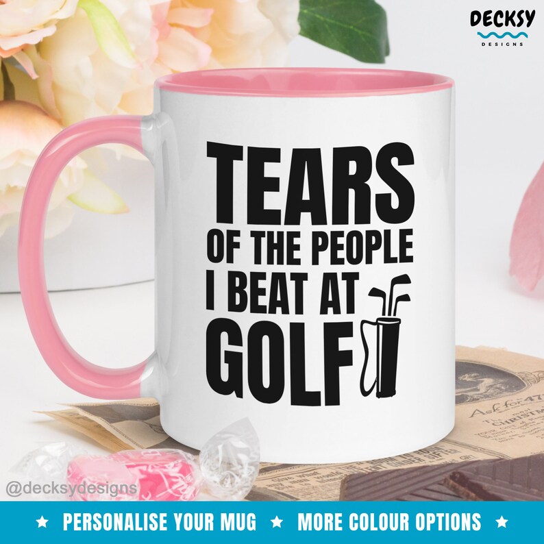 May include: White ceramic coffee mug with a pink rim. The mug has black text that reads "Tears of the people I beat at golf" with a golf bag and clubs graphic.
