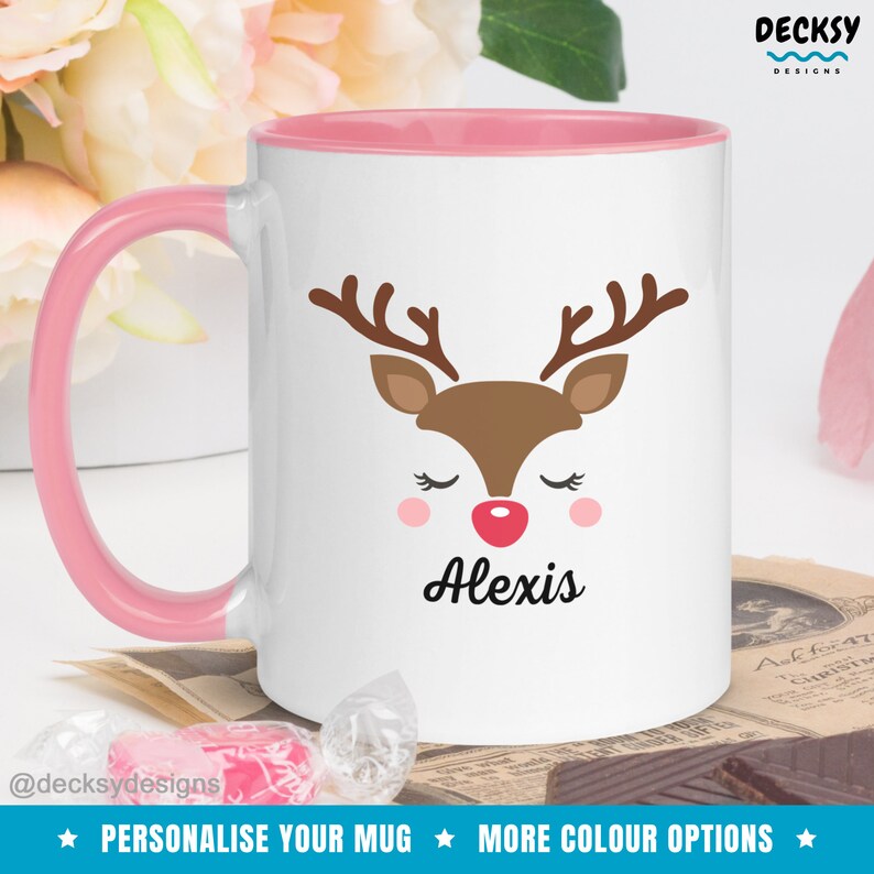 Op de afbeelding: Witte keramische mok met een roze rand en handvat. De mok heeft een cartoon rendiergezicht met gesloten ogen en een rode neus. De naam "Alexis" staat onder het rendier gedrukt.