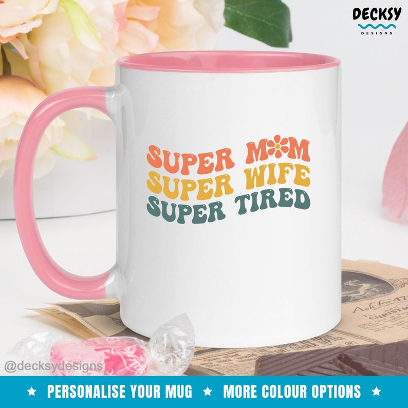 K&ouml;nnte beinhalten: Wei&szlig;e Keramiktasse mit rosafarbenem Rand. Die Tasse hat ein Retro-Design mit dem Text "SUPER MOM SUPER WIFE SUPER TIRED" in Regenbogenfarben.