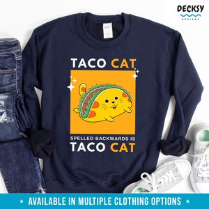 May include: A navy blue sweatshirt with the text "TACO CAT" in white letters. A cartoon cat wearing a taco is inside a yellow rectangle. The text "SPELLED BACKWARDS IS TACO CAT" is below the rectangle.