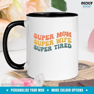 K&ouml;nnte beinhalten: Wei&szlig;e Keramiktasse mit schwarzem Rand. Die Tasse hat ein Retro-Design mit dem Text "SUPER MOM SUPER WIFE SUPER TIRED" in Regenbogenfarben.