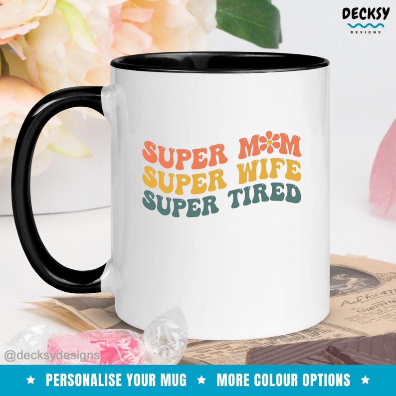 K&ouml;nnte beinhalten: Wei&szlig;e Keramiktasse mit schwarzem Rand. Die Tasse hat ein Retro-Design mit dem Text "SUPER MOM SUPER WIFE SUPER TIRED" in Regenbogenfarben.