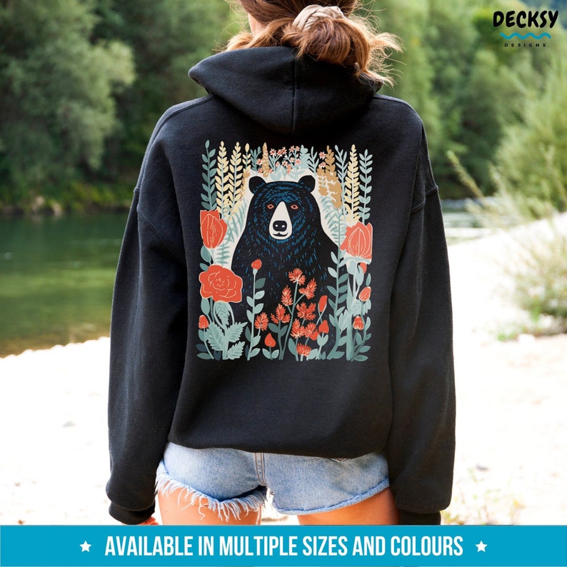 May include: Black hooded sweatshirt with a graphic print of a black bear in a field of flowers. The flowers are red, orange, and yellow. The text "DECKSY DESIGNS" is printed on the upper right corner of the sweatshirt.