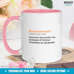 May include: White coffee mug with a pink rim and handle. The mug features the word "Booktrovert" defined as "Someone who prefers the company of fictional characters to real people."