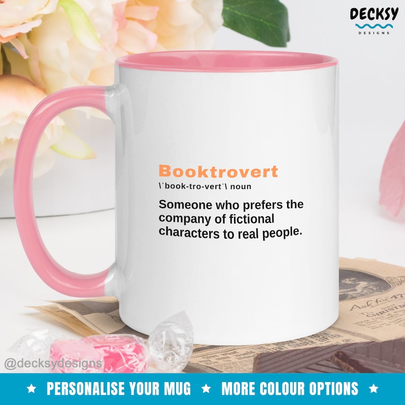 May include: White coffee mug with a pink rim and handle. The mug features the word "Booktrovert" defined as "Someone who prefers the company of fictional characters to real people."
