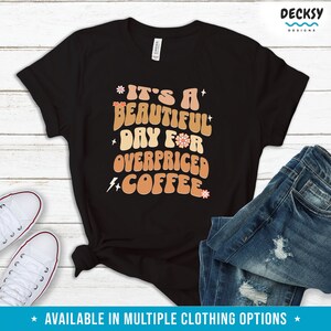 May include: Black t-shirt with a retro-style graphic that says "It's a beautiful day for overpriced coffee". The graphic is in orange, brown, and pink with a flower and lightning bolt design.