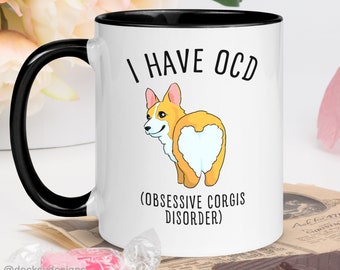 Może przedstawiać: Biały kubek ceramiczny z czarnym rantem i uchwytem. Kubek przedstawia kreskówkową grafikę tylnej części ciała corgi z sercowatą plamą białego futra. Na kubku nadrukowany jest tekst "I HAVE OCD (OBSESSIVE CORGIS DISORDER)".