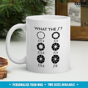 Könnte beinhalten: Eine weiße Keramiktasse mit einer schwarzen Grafik, auf der "WHAT THE f?" steht und eine Reihe von Blendenöffnungen für ein Kameraobjektiv dargestellt sind. Die Einstellungen sind f/1.4, f/2, f/2.8, f/4, f/5.6 und f/8.