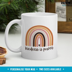 May include: White ceramic mug with a rainbow design in shades of brown, pink, and gray. The text "kindness is power" is printed below the rainbow. A small heart is in the center of the rainbow.