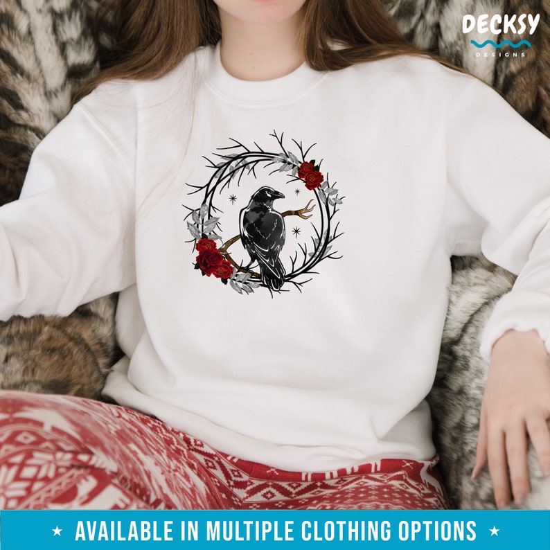 May include: White sweatshirt featuring a black raven perched on a branch within a wreath of dark branches, red roses, and gray leaves. The design includes small stars. The text at the bottom says "AVAILABLE IN MULTIPLE CLOTHING OPTIONS".