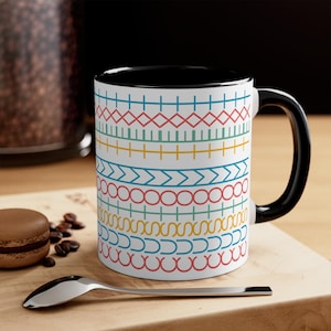 May include: White ceramic mug with a black interior and handle. The mug features a repeating pattern of colourful geometric shapes in red, blue, yellow, and green. A spoon and a macaron are in the foreground.