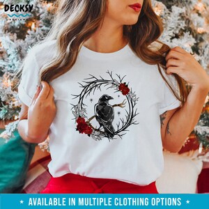 May include: White t-shirt featuring a black raven perched on a branch within a circular wreath of branches, red roses, and gray leaves. The text "AVAILABLE IN MULTIPLE CLOTHING OPTIONS" is at the bottom. The shirt has short sleeves.