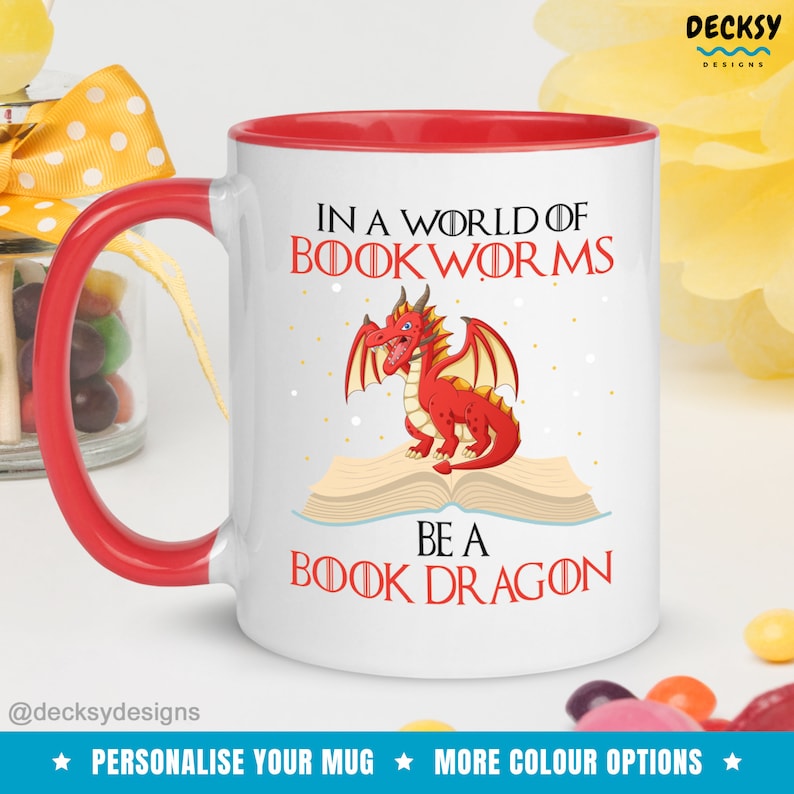 May include: White ceramic mug with a red rim and handle. The mug features a cartoon red dragon sitting on an open book. The text on the mug reads "In a world of bookworms be a book dragon".