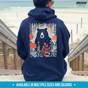 May include: Navy blue hoodie with a black bear surrounded by flowers and greenery on the back. The design is in a vintage style with red, orange, and green colors.