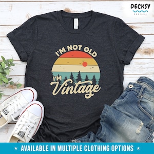 May include: A dark gray t-shirt with a vintage-style sunset graphic and the text "I'm not old, I'm vintage".