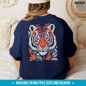 Puede incluir: Una sudadera azul marino con un gr&aacute;fico de una cabeza de tigre naranja y blanco rodeada de dise&ntilde;os florales naranja y blanco. El texto "DECKSY DESIGNS" est&aacute; en la esquina superior derecha de la imagen.