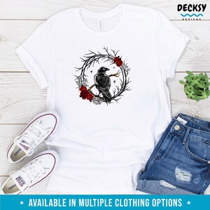 May include: White t-shirt featuring a black raven perched on a branch within a wreath of dark branches, red roses, and silver leaves. The text "AVAILABLE IN MULTIPLE CLOTHING OPTIONS" is at the bottom. The shirt is paired with denim shorts and white sneakers.