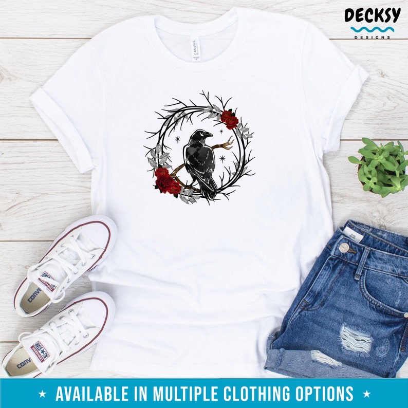 May include: White t-shirt featuring a black raven perched on a branch within a wreath of dark branches, red roses, and silver leaves. The text "AVAILABLE IN MULTIPLE CLOTHING OPTIONS" is at the bottom. The shirt is paired with denim shorts and white sneakers.