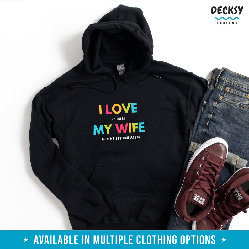 Pu&ograve; includere: Felpa con cappuccio nera con la scritta "I LOVE IT WHEN MY WIFE LETS ME BUY CAR PARTS" a colori. La felpa ha una tasca frontale e un cappuccio con coulisse. L'immagine mostra anche un paio di jeans e scarpe da ginnastica bordeaux.