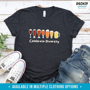 May include: Dark grey t-shirt featuring the phrase "Celebrate Diversity" above a line of beer glasses in different colours. White trainers and blue jeans are also visible. The shirt is part of a clothing range.