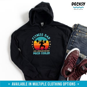 Puede incluir: Sudadera con capucha negra con un gr&aacute;fico de estilo vintage que dice "Fitness Dad" con una silueta de una persona levantando pesas. El texto debajo del gr&aacute;fico dice "Como un padre normal pero mucho m&aacute;s cool."