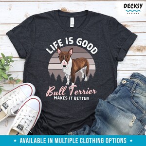 May include: A dark gray t-shirt with a white and pink graphic design. The graphic features a brown and white bull terrier dog standing in front of a sunset with the text "Life is Good" above the dog and "Bull Terrier Makes it Better" below the dog.