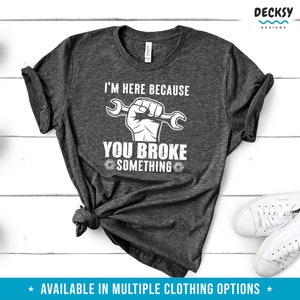 Puede incluir: Una camiseta gris oscuro con el texto "I'm here because you broke something" en blanco. Una ilustraci&oacute;n blanca de un pu&ntilde;o sosteniendo una llave inglesa est&aacute; por encima del texto.