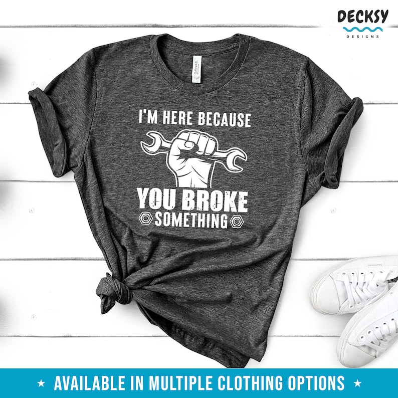 Puede incluir: Una camiseta gris oscuro con el texto "I'm here because you broke something" en blanco. Una ilustraci&oacute;n blanca de un pu&ntilde;o sosteniendo una llave inglesa est&aacute; por encima del texto.