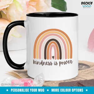 May include: White ceramic mug with a black rim and handle. The mug features a rainbow design with a brown heart in the center and the text "kindness is power".