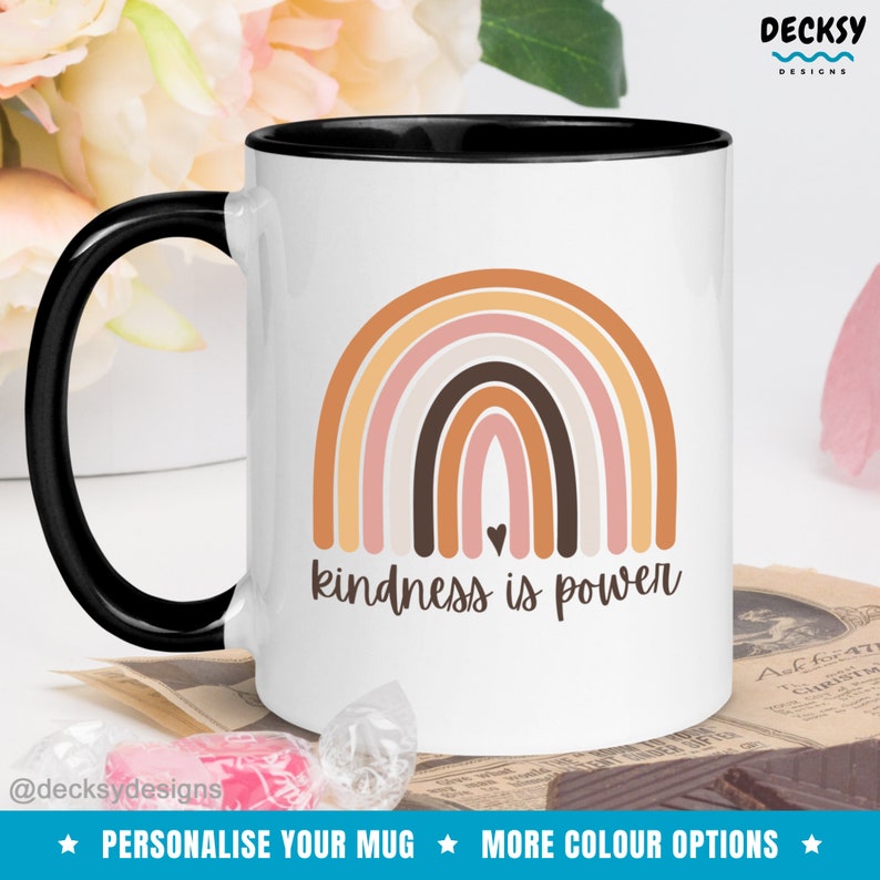 May include: White ceramic mug with a black rim and handle. The mug features a rainbow design with a brown heart in the center and the text "kindness is power".