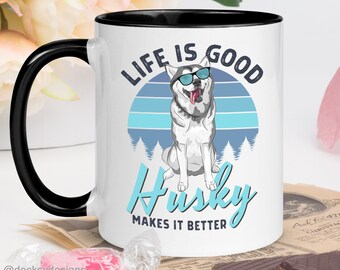 Puede incluir: Taza de cerámica blanca con borde y asa negros. La taza presenta un dibujo animado de husky con gafas de sol y un diseño de puesta de sol retro. El texto "Life is good Husky makes it better" está impreso en la taza.