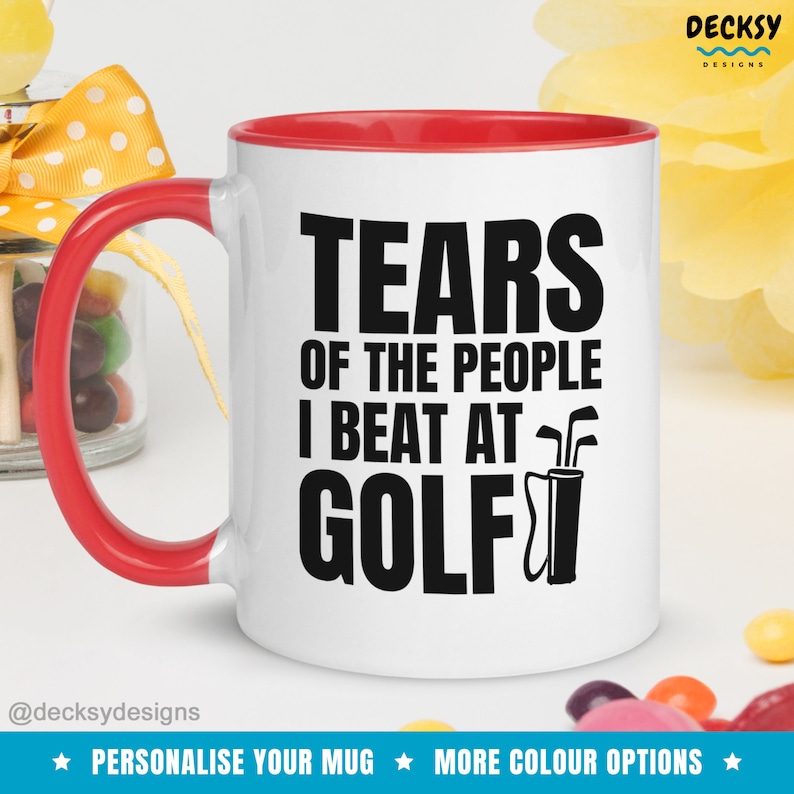 May include: White ceramic coffee mug with a red rim. The mug has a black graphic that says "Tears of the people I beat at golf" with a golf bag and clubs.