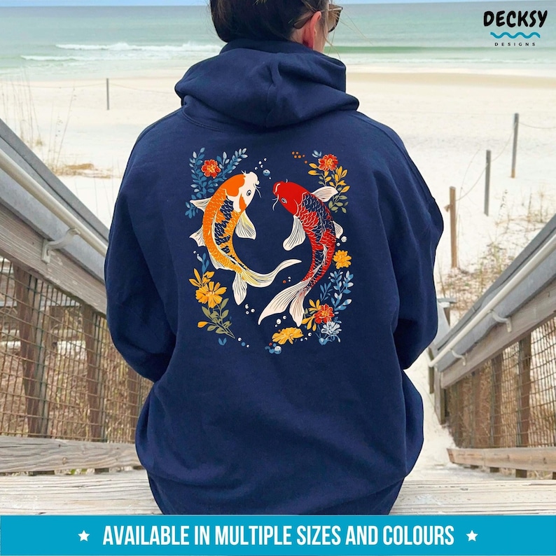 May include: A navy blue hoodie featuring a design of two koi fish swimming in a circle of flowers. The fish are orange and red with white scales. The flowers are yellow, orange, and blue.