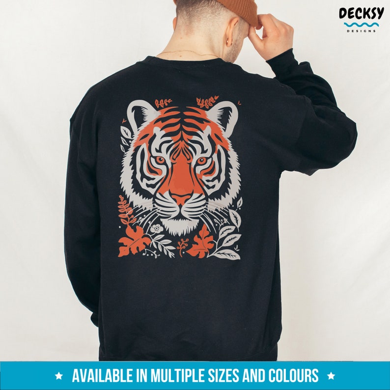 Puede incluir: Sudadera negra con un estampado gr&aacute;fico de la cara de un tigre con rayas naranjas y blancas, rodeado de hojas y flores. El texto "DECKSY DESIGNS" est&aacute; en la esquina superior derecha de la imagen. El texto "AVAILABLE IN MULTIPLE SIZES AND COLORS" est&aacute; en la parte inferior de la imagen.