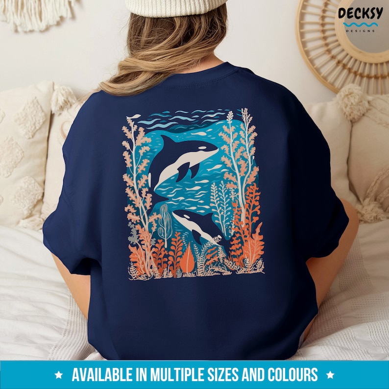 May include: A navy blue t-shirt with a graphic of two orcas swimming in a coral reef. The orcas are black and white, and the coral reef is a mix of orange, pink, and blue.