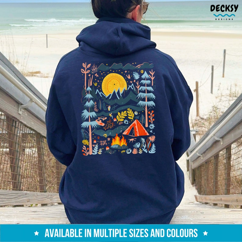 May include: A navy blue hoodie with a colorful illustration of a campsite scene on the back. The illustration features a tent, a campfire, trees, mountains, and a full moon. The text "DECKSY DESIGNS" is printed in white on the upper right corner of the hoodie.