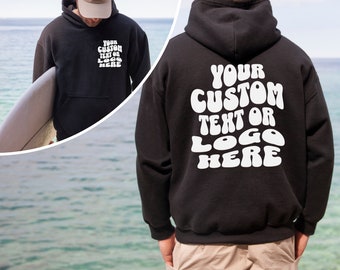 May include: Black hooded sweatshirt with white text that reads "YOUR CUSTOM TEXT OR LOGO HERE" in a retro font. The sweatshirt is shown on a person standing on a beach.
